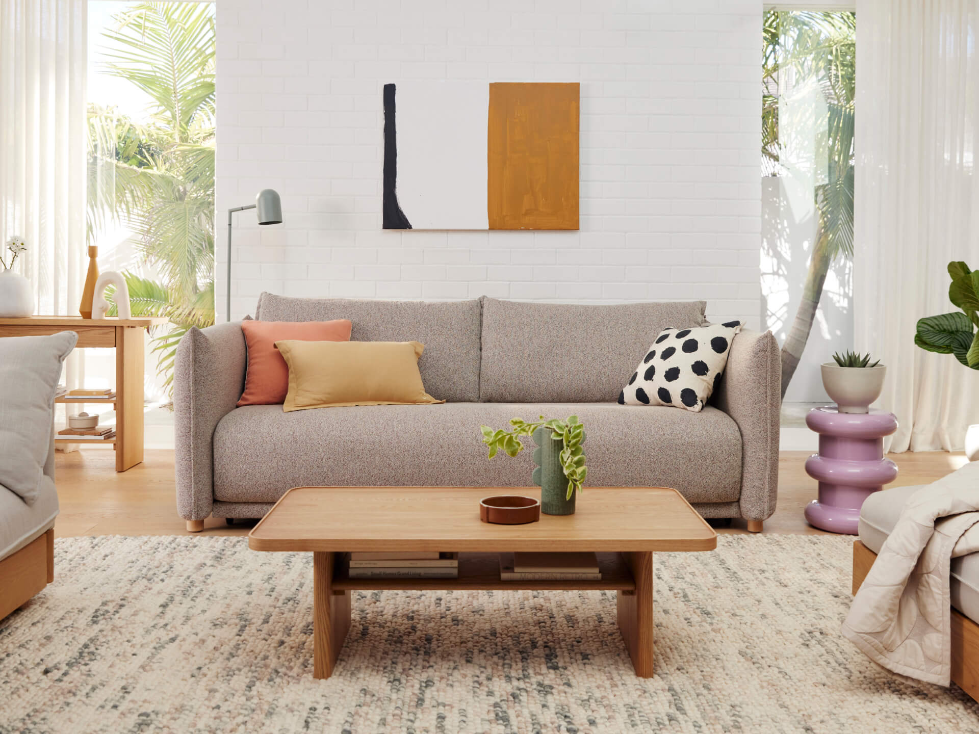 A light grey sofa with colorful cushions sits in a bright room beside a wooden coffee table, a pink side table, and green plants.