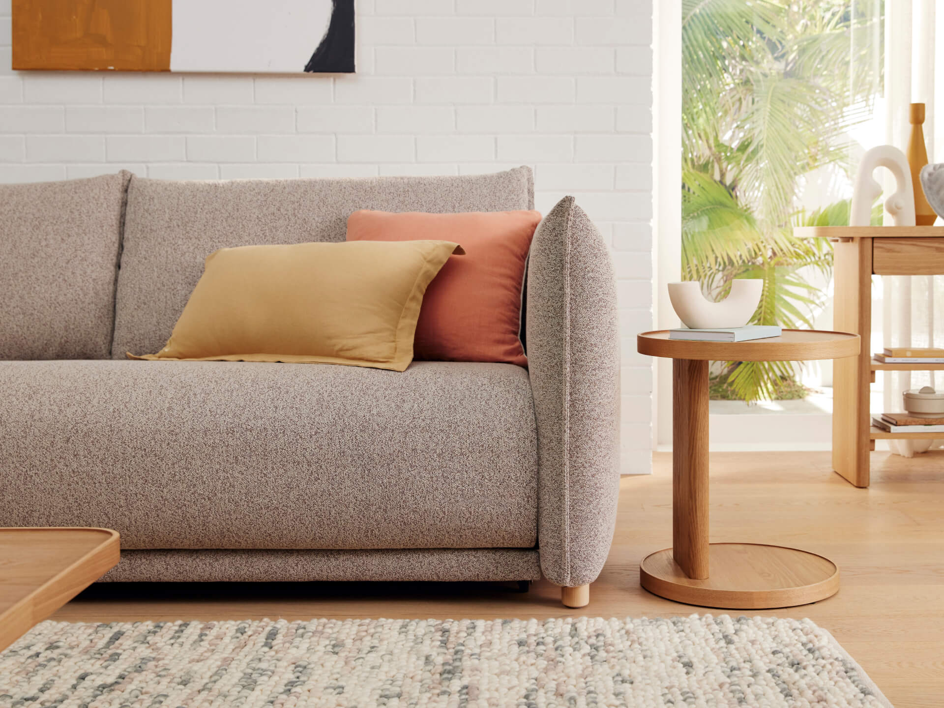 A light grey textured sofa with a mustard yellow and coral pink pillow, next to a round wooden side table on a light colored rug.