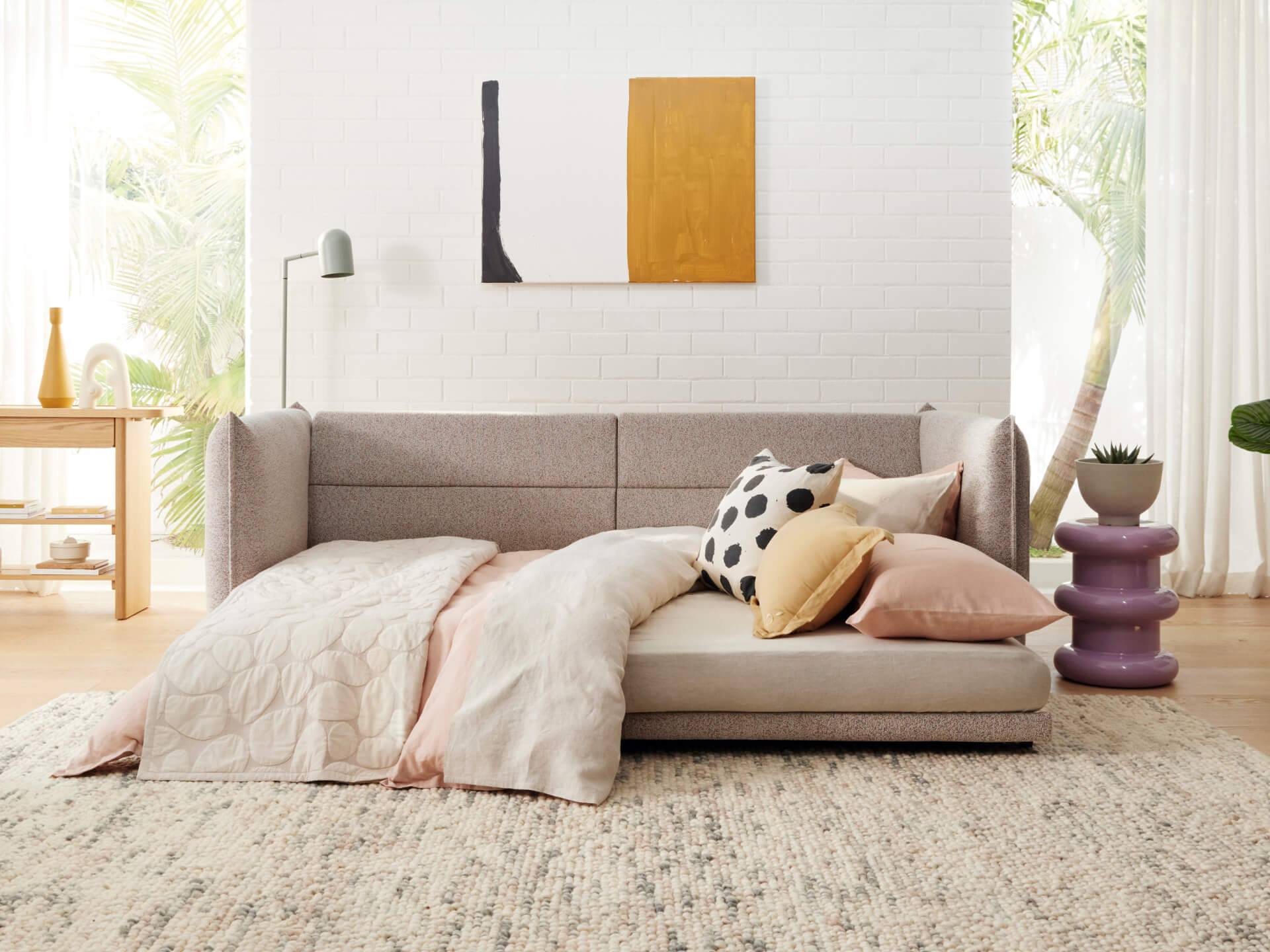 A light grey sofa with colorful cushions, a pale pink throw, and a purple side table, beside a wood table and a vibrant abstract painting.