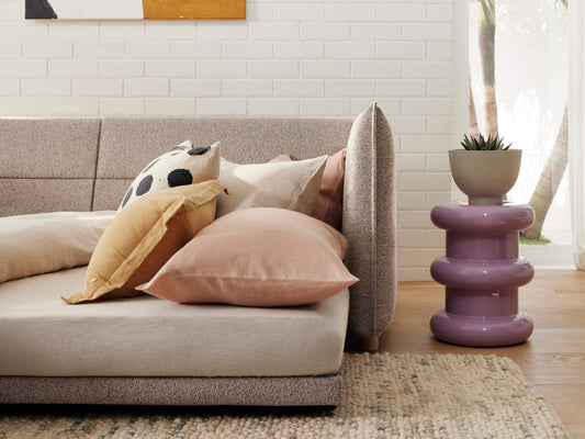 A light beige sofa with pastel pillows, including a tan and a blush, beside a shiny purple side table holding a gray pot with a plant.