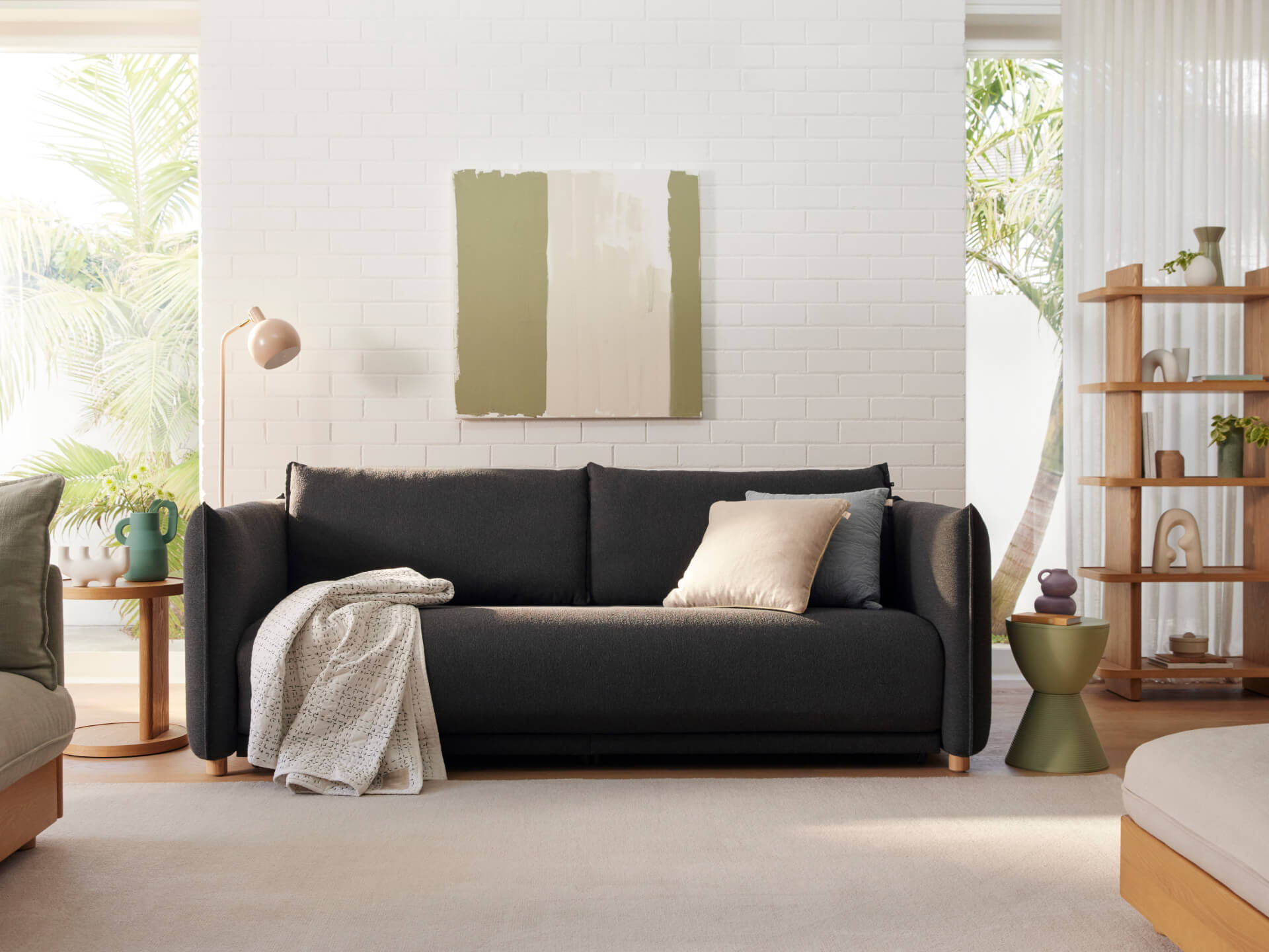 A dark grey sofa with a beige pillow and white blanket, beside a light wood table, green vase, and an olive green accent table.