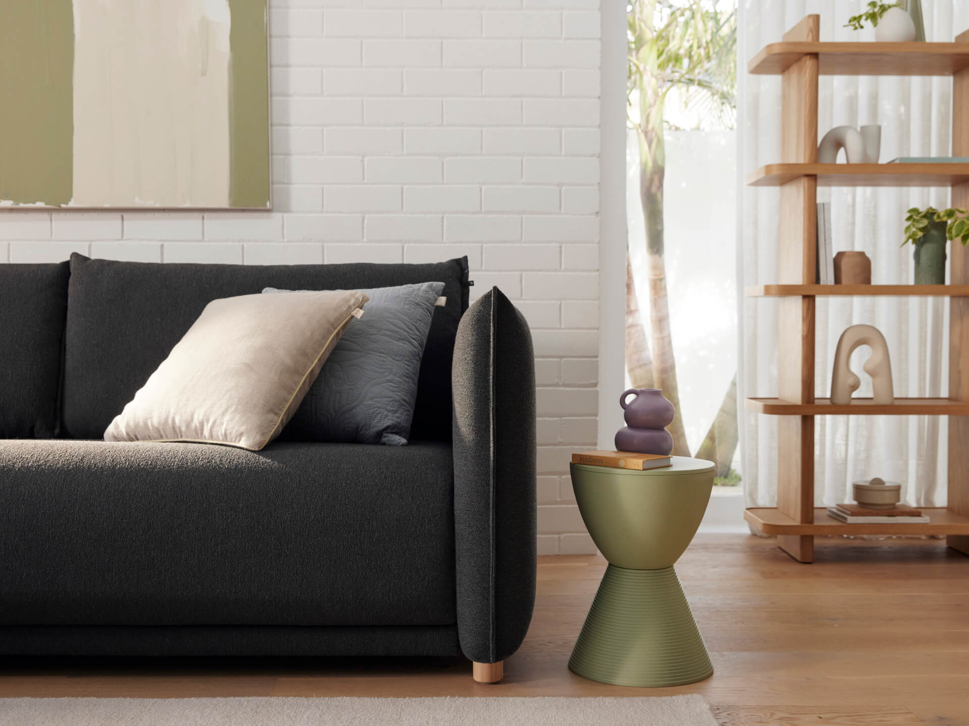 A dark grey sofa with a beige and a blue cushion beside a green accent table holding a purple vase, against a white brick wall.