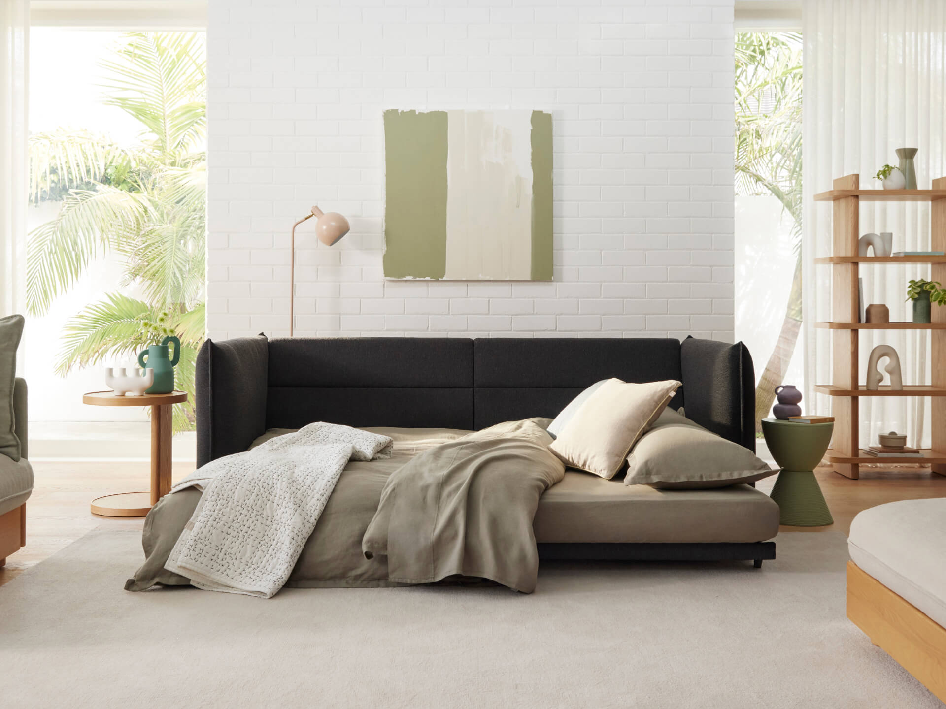 A dark grey sofa with pillows lies on a light beige rug. A green throw blanket drapes over it. Nearby are a wooden side table and a floor lamp.