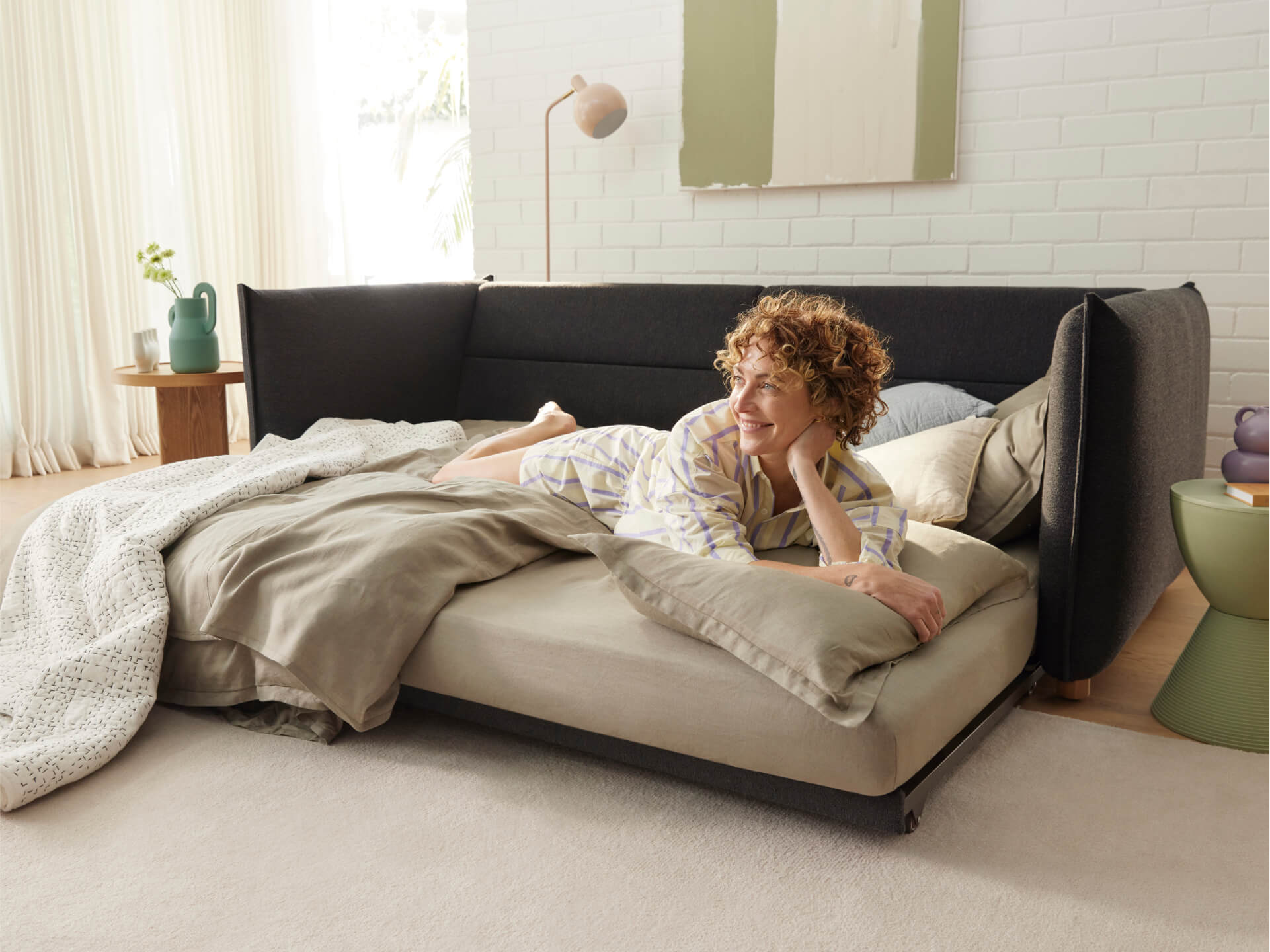 A woman with curly hair relaxes on a beige bed with light brown bedding, beside a green side table and a jar in soft green tones.
