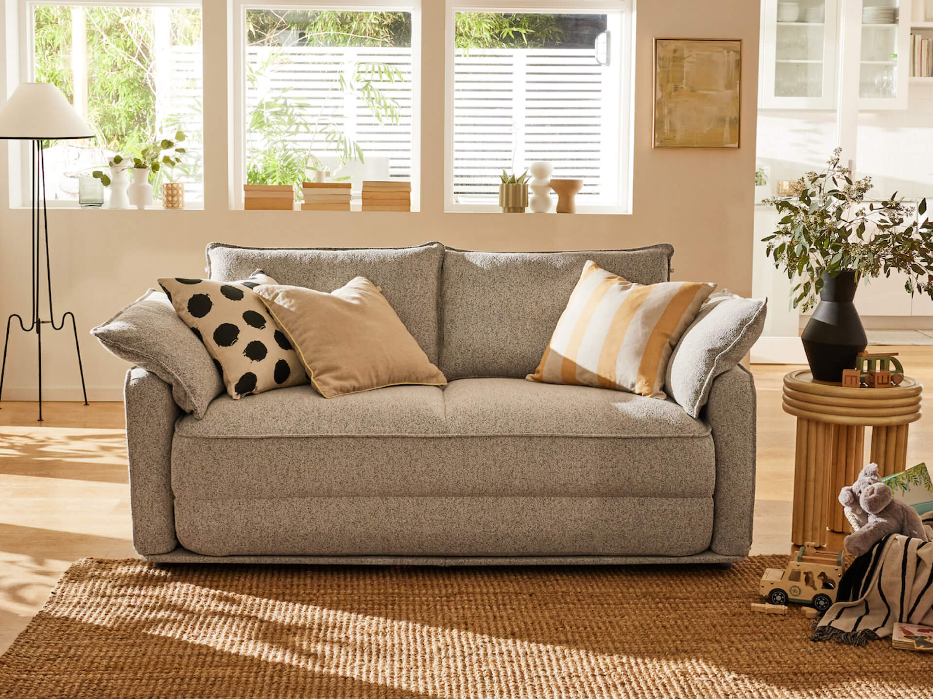 A light grey sofa with cushions; one beige with black dots, another striped in gold and white. A woven rug, potted plants, and a lamp are visible.