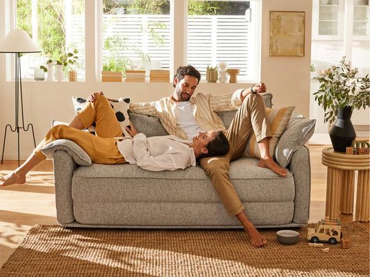 A couple relaxes on a light grey sofa with textured fabric, surrounded by plants, wood accents, and natural light in a warm living room.