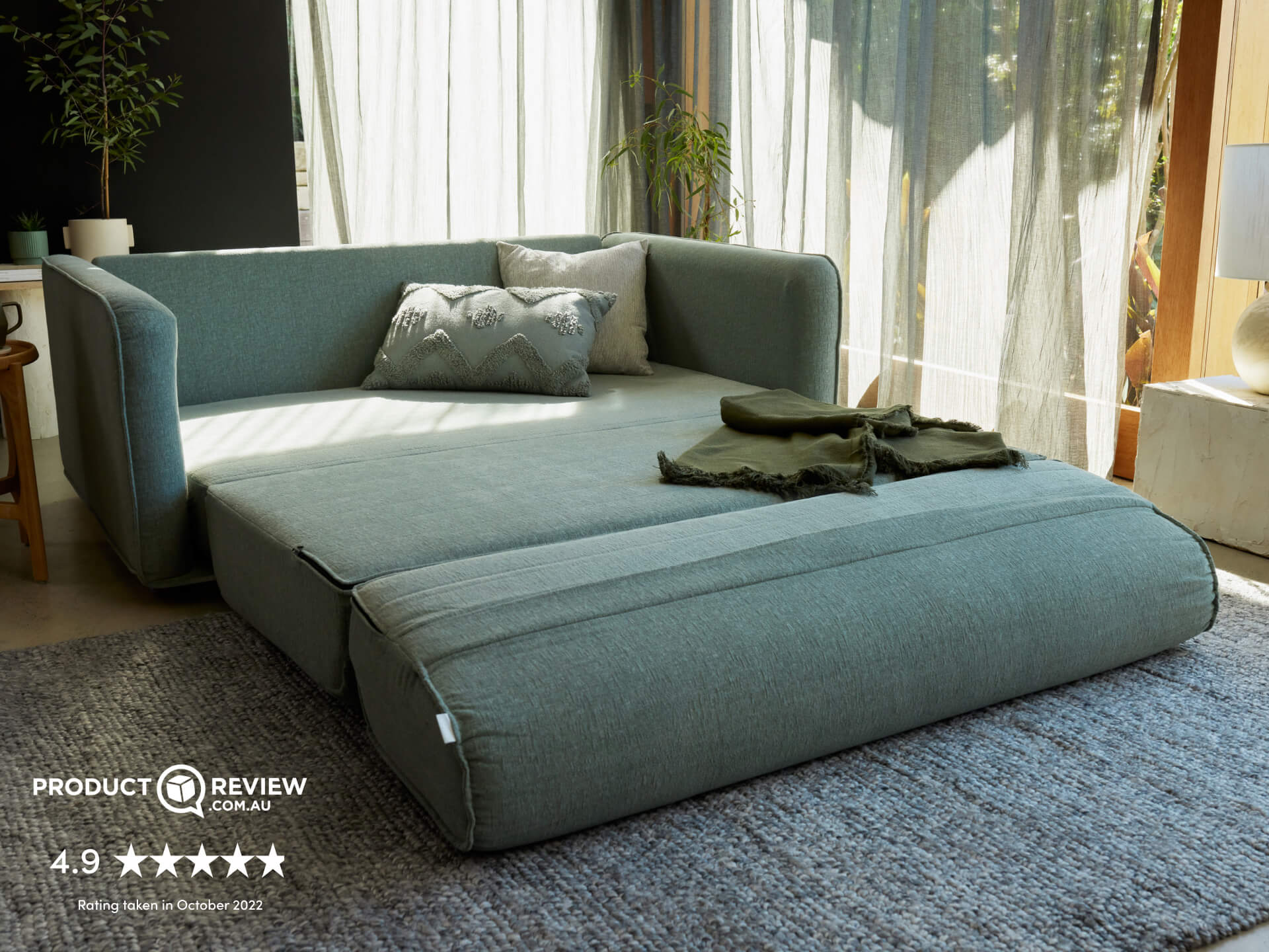 A light teal modular sofa with a removable cushion resting on a textured grey rug, beside sheer curtains and a potted plant.