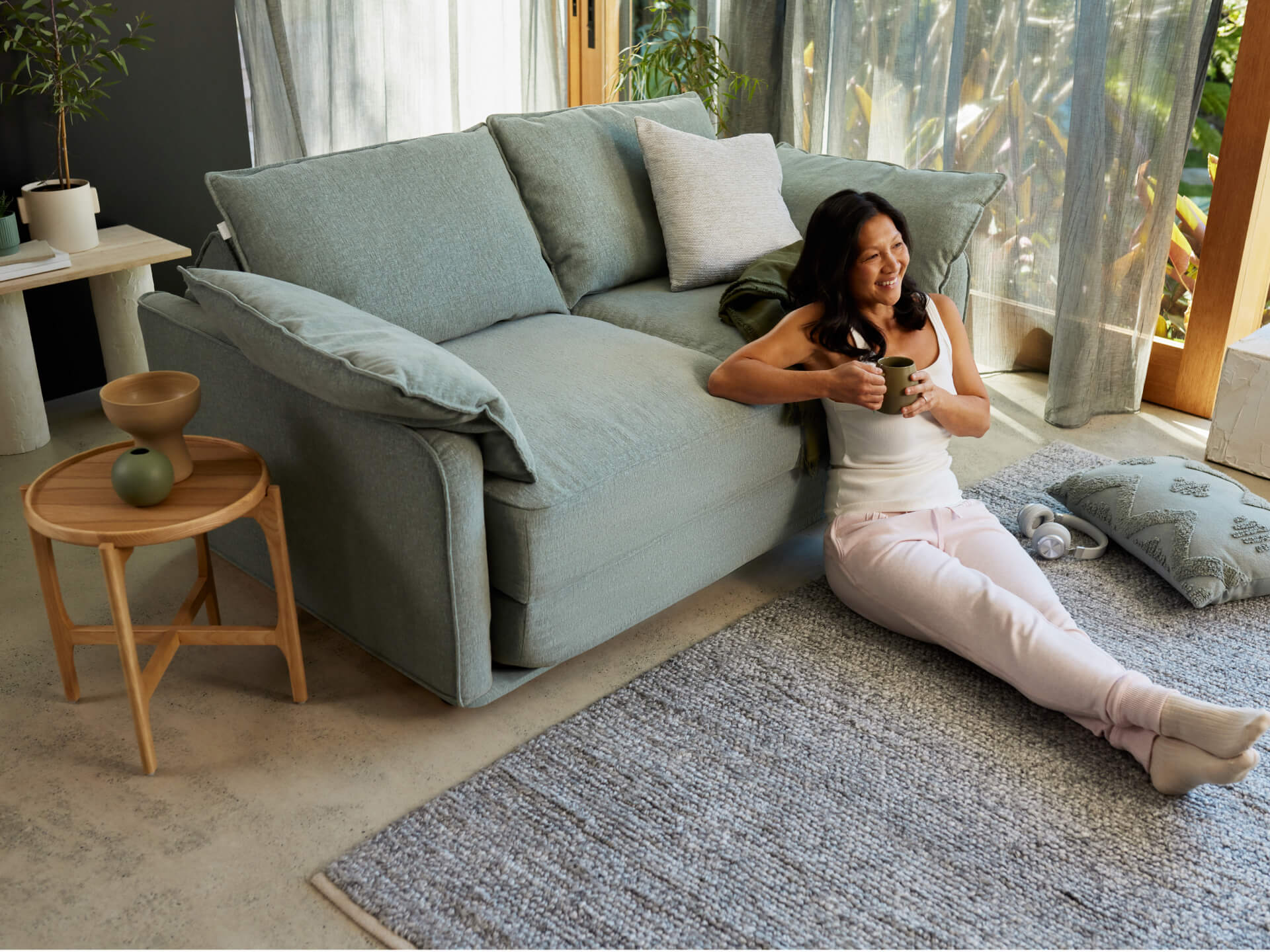A woman in a white tank top sits on a light grey sofa, holding a green mug. She wears light pink sweatpants, with a soft grey rug below.