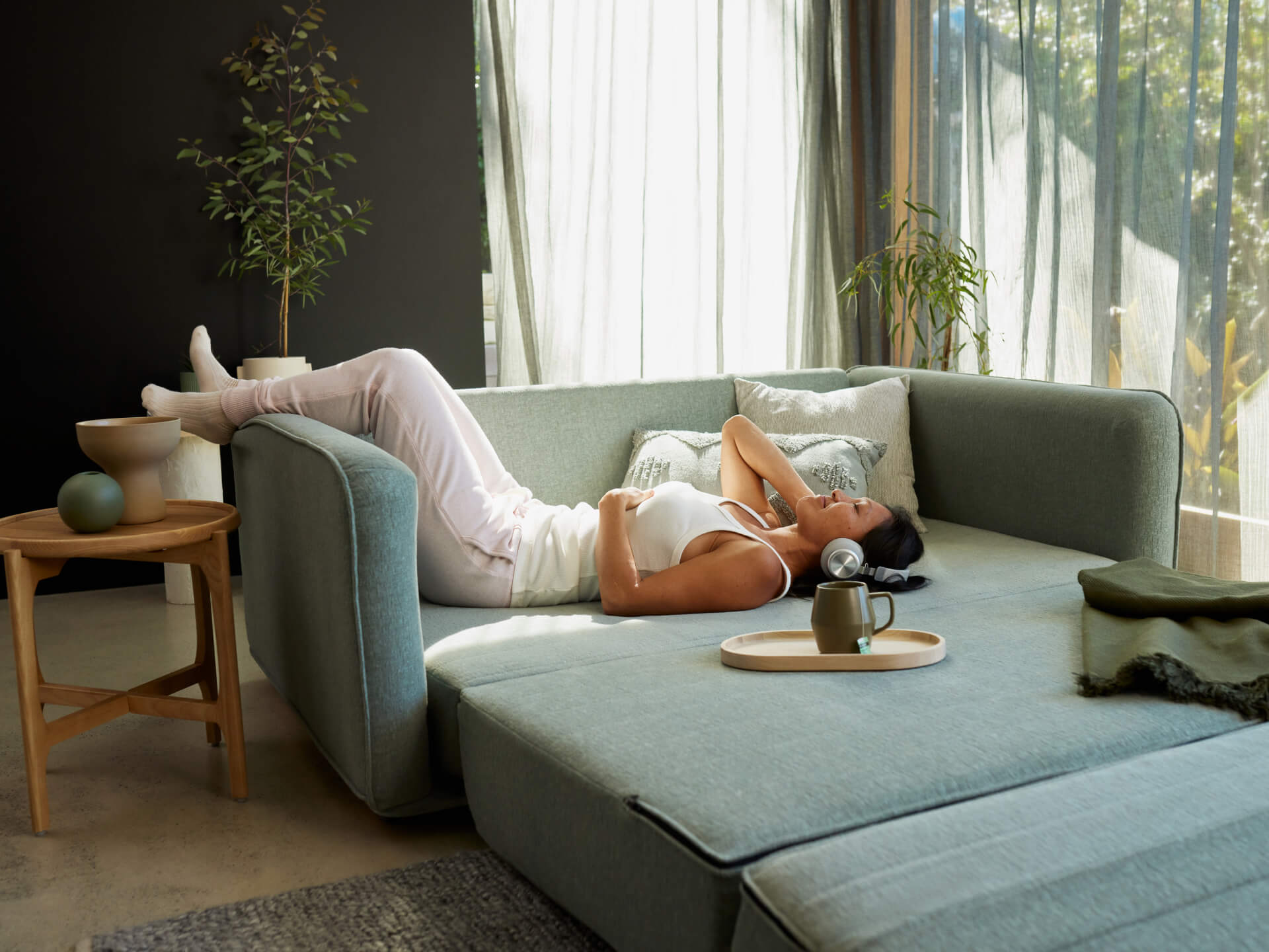 A woman in a white tank top lies on a light teal sofa, with pale pink pants and white socks. A wooden table holds green and beige decor.