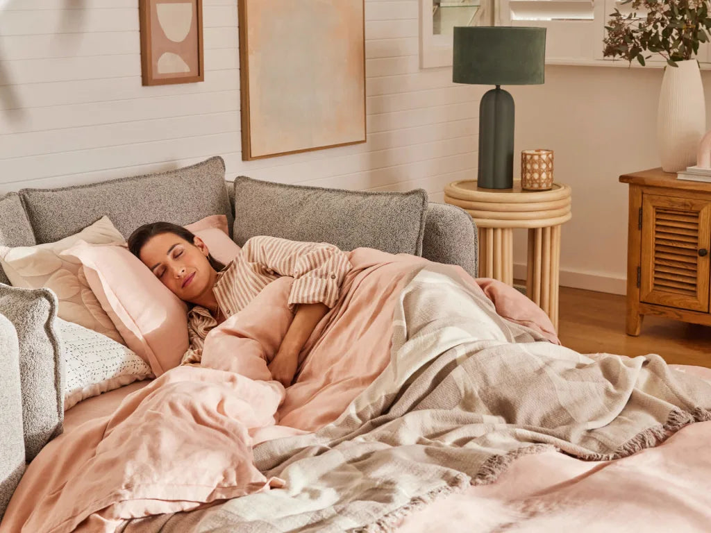 A woman with brown hair sleeps peacefully on a light grey sofa, surrounded by soft pink and beige blankets and pillows. A green lamp and wooden table are nearby.