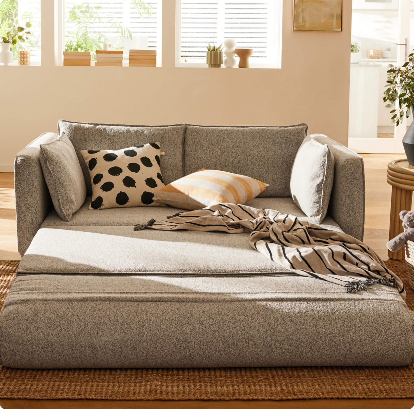 A light grey sofa with a black polka dot pillow and a mustard-striped pillow. A beige striped throw drapes over the seat, set on a woven rug.