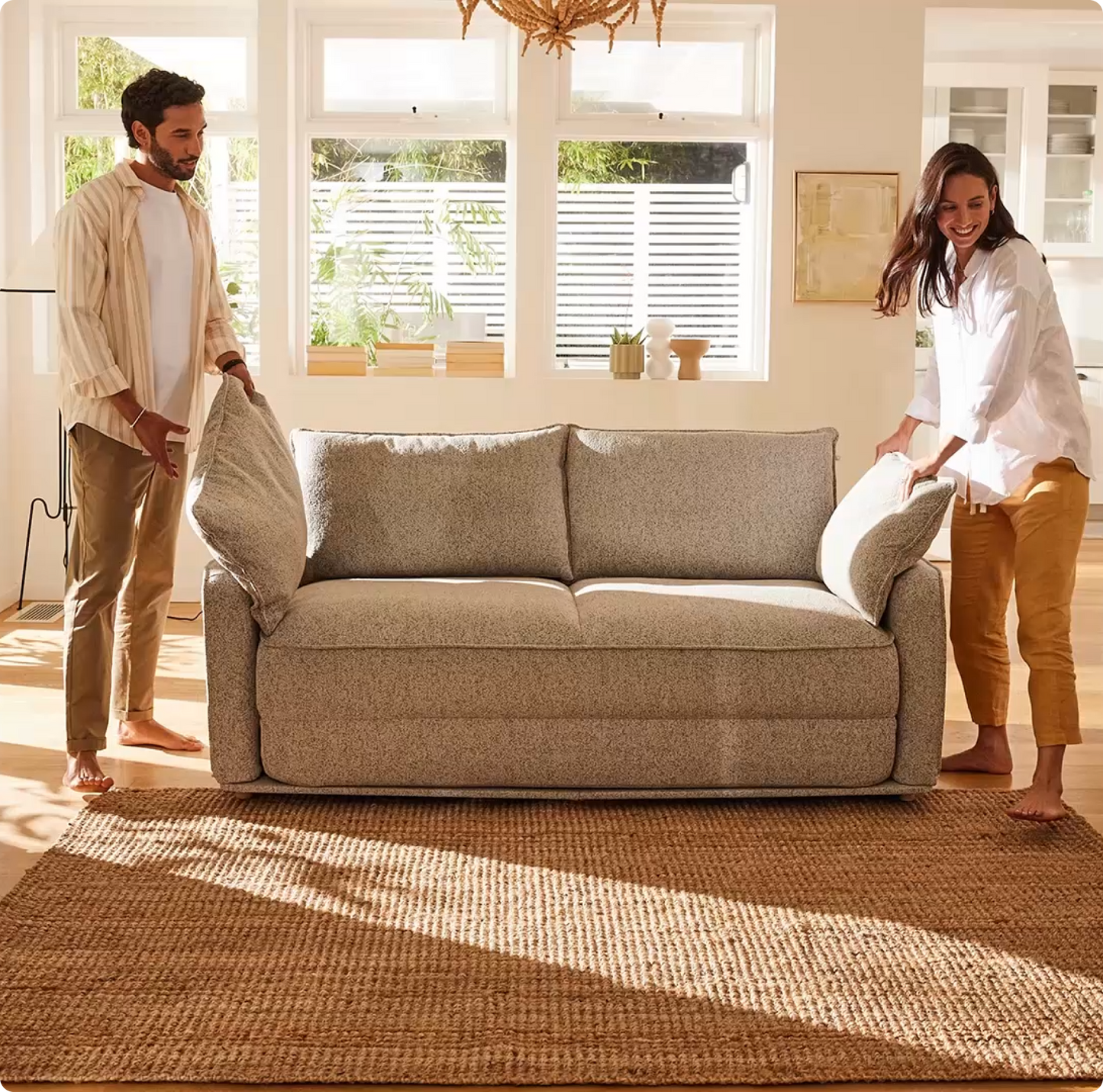 A couple rearranges a light grey sofa with textured cushions on a jute rug, in a bright room with large windows and natural light.