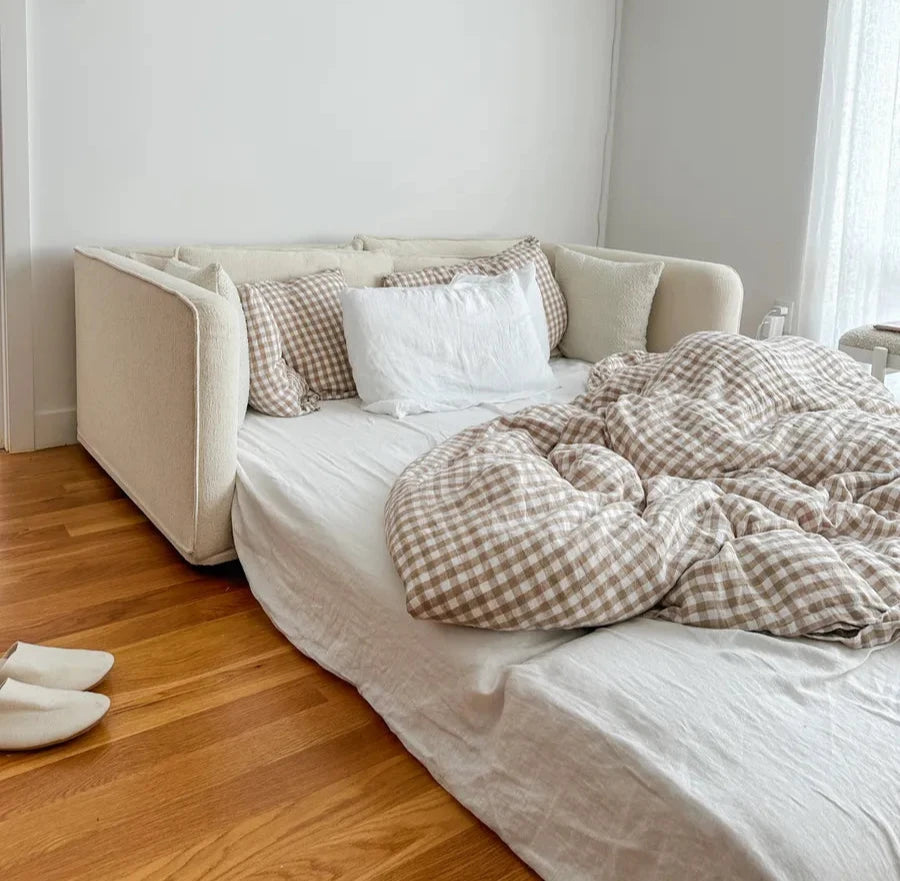 A light beige sofa with a bed setup; white and brown checkered bedding is rumpled. White pillows sit against a plain wall. Hardwood floor visible.