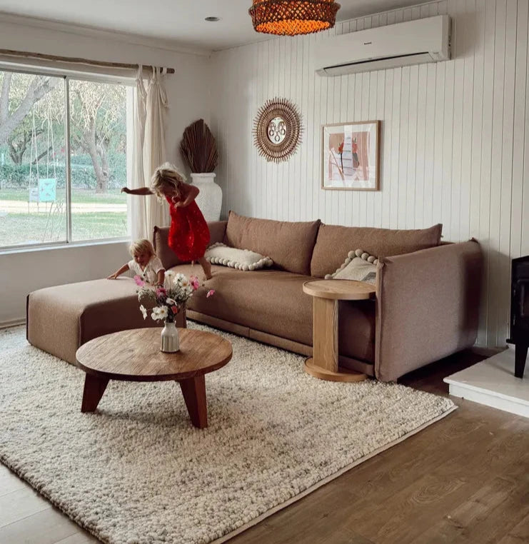 Cozy living room with a brown sectional sofa, a round wood coffee table, and playful children. A white rug and cream walls create a warm atmosphere.