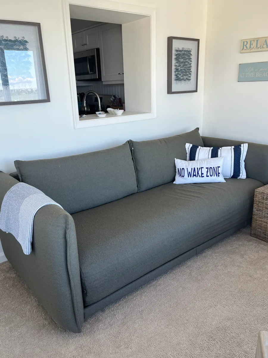 A dark green sofa with a light grey throw blanket, featuring a white pillow with navy stripes and text, against a beige carpet.